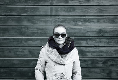 Young woman wearing winter coat and sunglasses against wooden wall