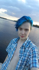 Portrait of young man standing against lake with blue dyed hair