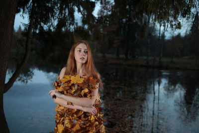 Woman wearing dress made of autumn leaves while standing against lake at dusk