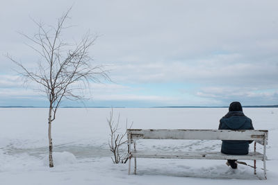 Rear view of man sitting on bench in snow-coverd landscape against sky