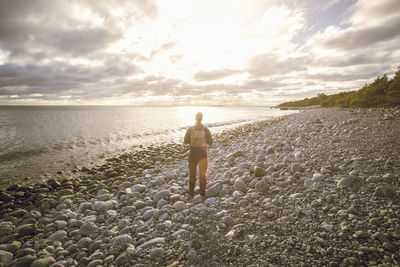 Rear view of man standing on rocky shore at beach against sky during sunset