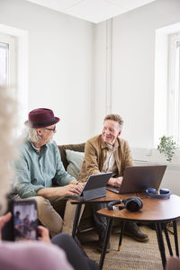 Senior men talking to each other while sitting in living room and using digital tablet and laptop to edit podcast