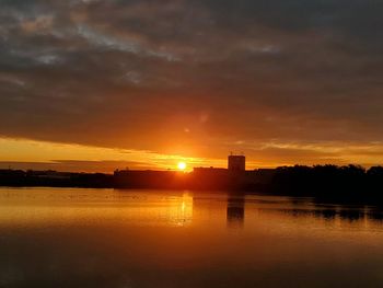 Silhouette buildings by lake against sky during sunset