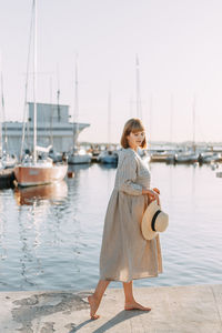 Happy young woman in dress and hat walks on the beach near the boats