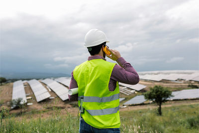 Back view of unrecognizable male holding blueprint while speaking on walkie talkie against solar power station and overcast gray sky