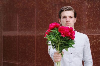 Portrait of handsome man standing against red wall holding flower in hand