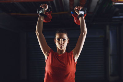 Waist up portrait of young woman with arms raised holding kettlebells