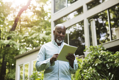 Senior man looking at tablet outside house on sunny day