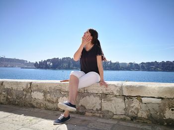 Mature woman sitting on retaining wall by river against clear blue sky