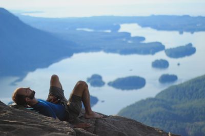 Man sitting on rock by lake against sky