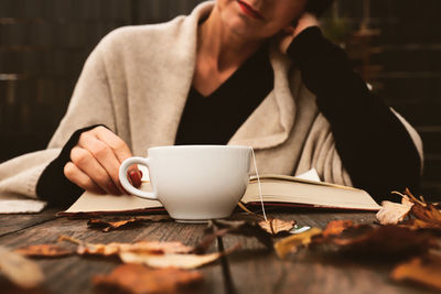 Midsection of woman reading book by tea cup on table