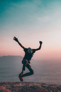 Full length of man jumping on top of a cliff against mountain and sky during sunset