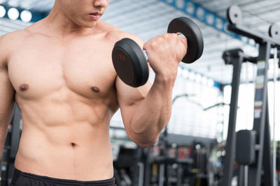 Midsection of shirtless man lifting dumbbell at gym