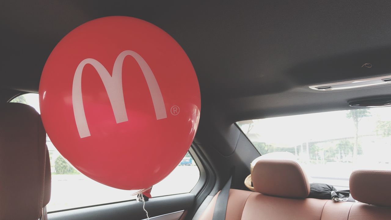 vehicle interior, mode of transportation, communication, transportation, red, land vehicle, car, text, motor vehicle, real people, sign, day, car interior, indoors, close-up, glass - material, western script, window, one person, balloon