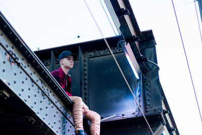 Low angle view of teenager boy sitting on railway bridge against clear sky