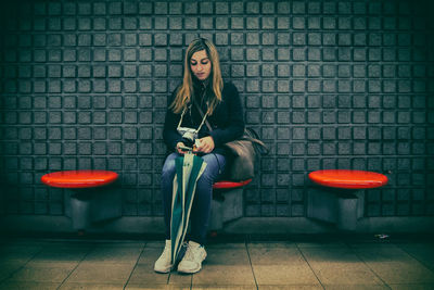 Full length of young woman using phone on seat against wall