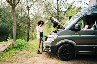 Young african american female traveler standing near camper van with opened hood and talking with repair service by mobile phone while getting troubles during summer trip in nature