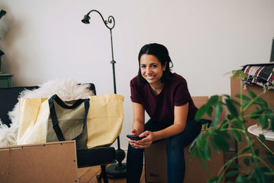 Portrait of smiling woman holding mobile phone while sitting on box against wall