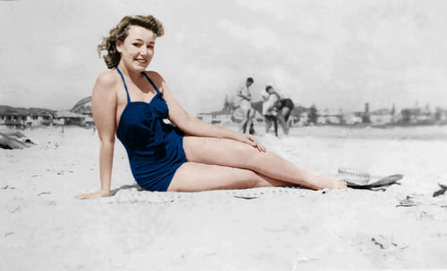 Portrait of young beautiful woman in dress sitting on sand against cloudy sky