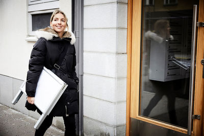 Confident female real estate agent with blank signboard walking on sidewalk in city