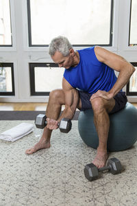 Full length of muscular mature man lifting dumbbell while sitting on fitness ball at home