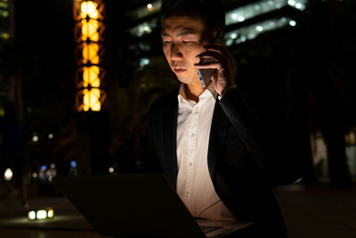 Focused asian male entrepreneur in formal wear browsing on laptop while having phone call sitting on street with glowing buildings in night time