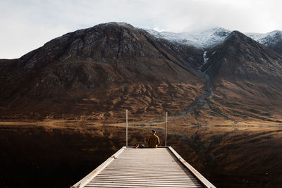 Distant view of lonely tourist sitting on wooden pier and admiring peaceful scenery of mountains and lake on overcast day in scottish highlands