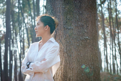 Thoughtful young woman standing by tree trunk in forest