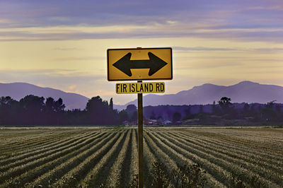 Road sign by agricultural field against sky during sunset