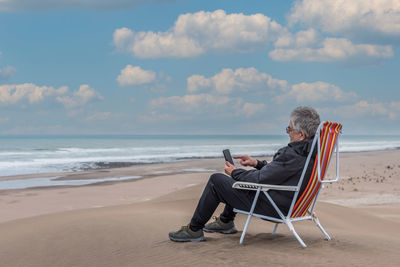 Adult man sitting on a beach chair on top of a beachfront medano typing on smartphone.