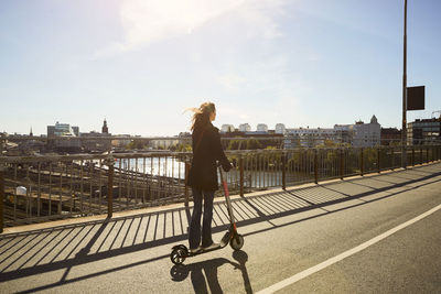 Full length of female commuter riding electric push scooter on bridge in city against sky