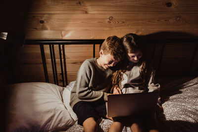 Smiling brother and sister using laptop while sitting on bed against wall at home