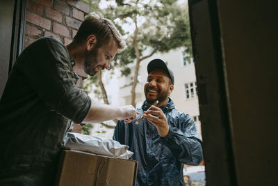 Smiling delivery person taking electronic signature from man while delivering packages