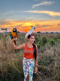 Rear view of girl standing on field against sky with an macaw and an cockatoo during sunset