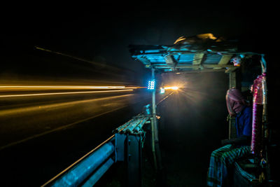 Man sitting at tea stall by light trails at night