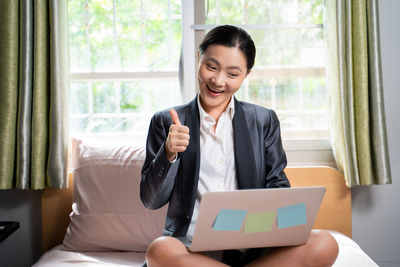 Young woman using mobile phone while sitting on window