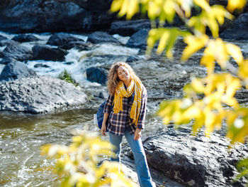 Young curly woman traveller smiling at mountain river in autumn