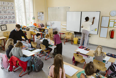 Male and female teacher teaching pupils sitting at desk in classroom