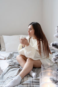 Woman in warm white winter sweater lying in bed at home at christmas eve holding cup