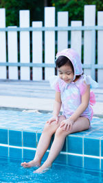 Charming 4 years old asian little girl wear swimsuit sitting happily at swimming pool outdoors.