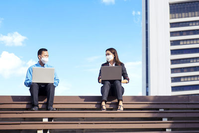 Business persons wearing mask using laptop sitting outdoors