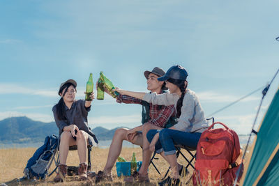 Happy friends toasting drinks while sitting on chairs against blue sky during sunny day