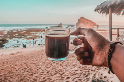 Cropped hand holding coffee at beach against sky during sunset
