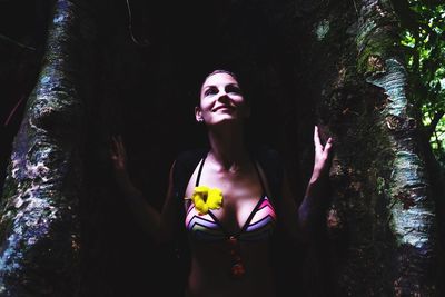 Smiling young woman in bikini looking up while standing by tree in forest