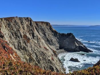 People so small in contrast to ocean cliffs against blue background.