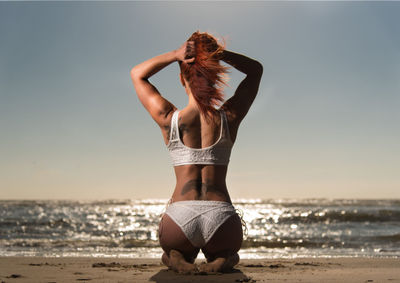 Rear view of sensuous female model kneeling in bikini at beach