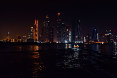 Illuminated buildings by river against sky at night