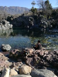 Rear view of man sitting on rock by lake