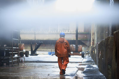 Rear view of manual worker wearing raincoat and helmet while walking at factory