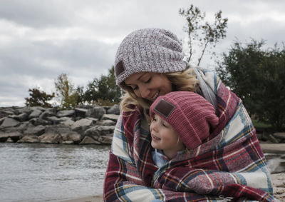 Happy mother with daughter wrapped in blanket sitting at beach against cloudy sky